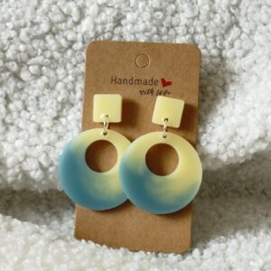 Paire de boucles d'oreilles en résine époxy en forme de rond creux surmontées d'un carré au niveau de l'attache, attachées à un carton fait main
