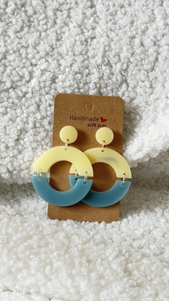 Paire de boucles d'oreilles en résine époxy en forme de deux demi-cercles attachés et surmontés d'un rond au niveau de l'attache, attachées à un carton fait main