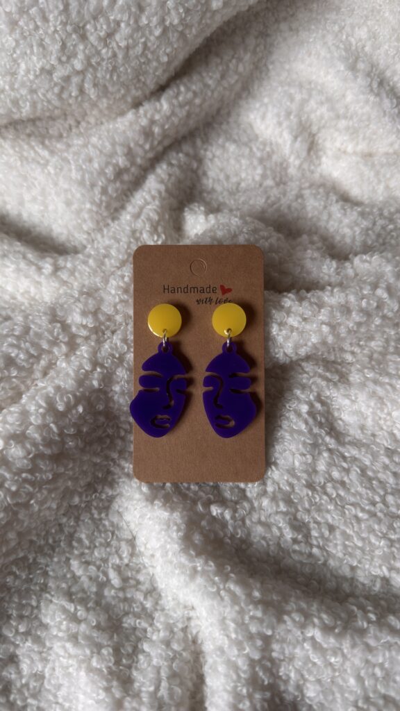 Boucles d'oreilles en résine époxy violette, en forme de silhouette de femme ajourée façon line art, surmontée d'une puce ronde jaune au niveau de l'attache. Attache en acier inoxydable.